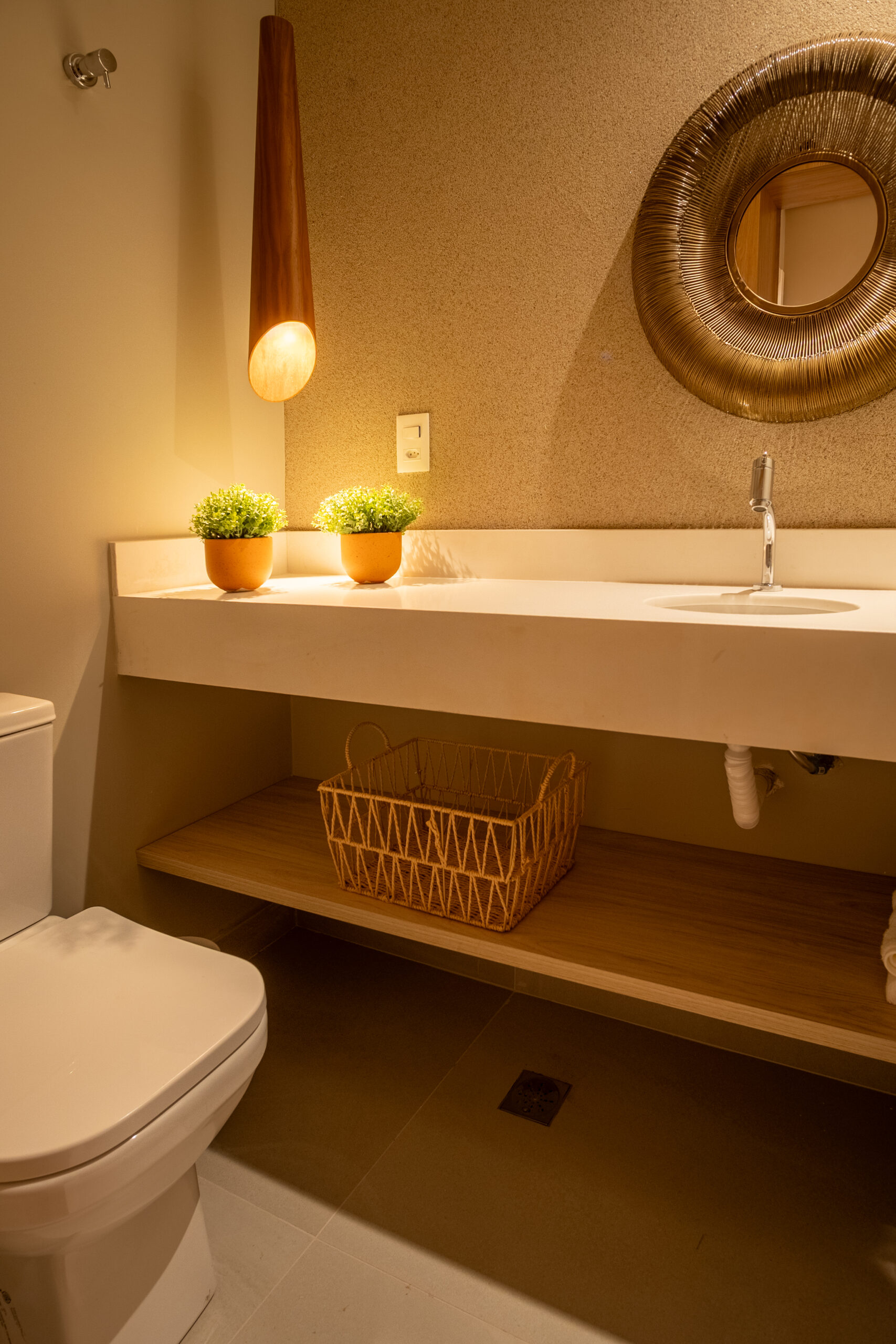 Banheiro moderno com bancada branca, iluminação pendente em madeira, espelho decorativo redondo e detalhes em tons naturais, como plantas e cesta de vime.